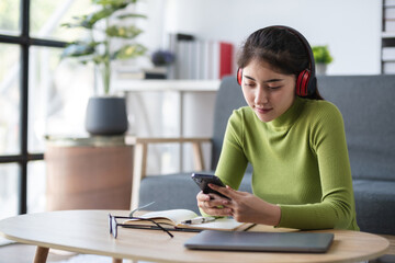 Asian woman using smartphone while working with headphones in modern office, concept of multitasking