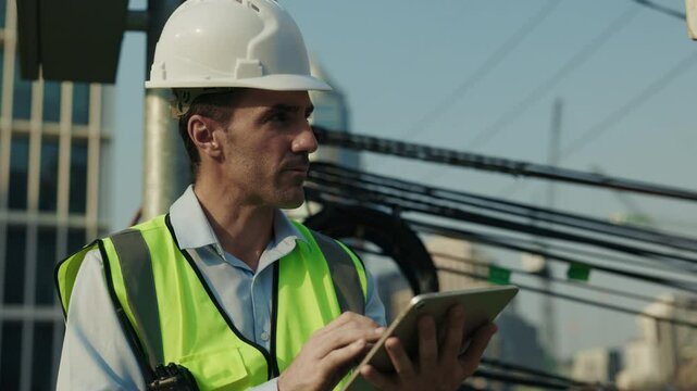 Portrait of man engineer in helmet working on tablet at construction site. Male supervisor in safety uniform compares work results with architectural documents looking away at sunset