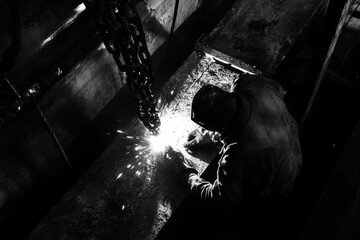 Industrial worker welding metal in a dark environment with bright sparks flying, showcasing craftsmanship and precision in a black and white composition
