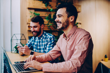 Cheerful male student laughing at funny joke learning on laptop computer spending time in campus with friend,happy prosperous freelancers satisfied with productive job using modern technologies