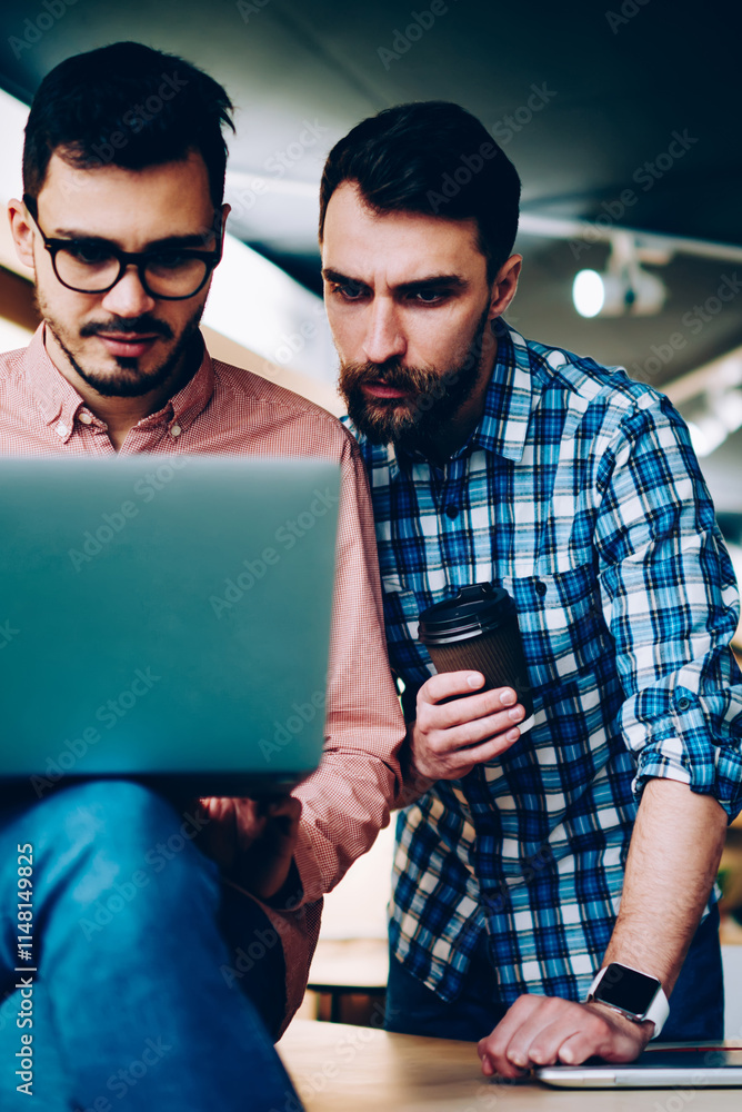 Poster serious male coworkers concentrated on fixing bugs of application on laptop computer looking togethe