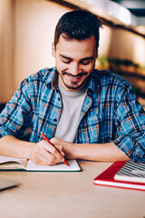 Smiling male blogger satisfied with having great idea for publication making notes sitting at desktop with netbook, positive student enjoying learning process doing homework task in college library