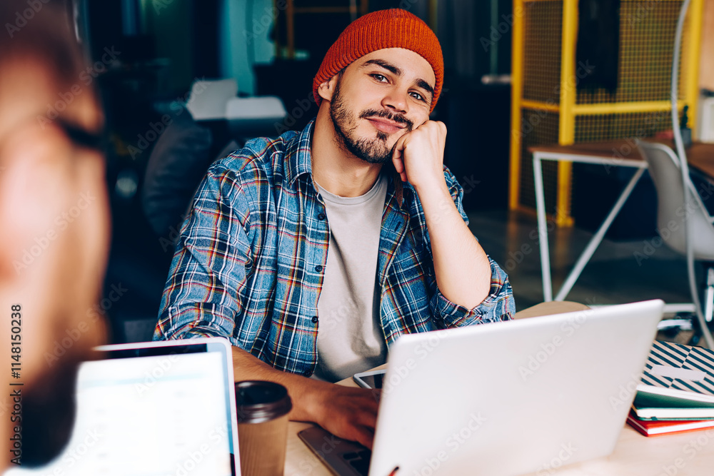 Poster contemplative hipster guy looking at camera while sitting at laptop computer for learning in college