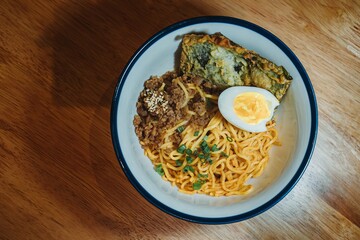 Noodles on a bowl with toppings: Half boiled egg, fried nori, a pile of minced chicken meat, green onion, and sesame. Flat lay, overhead, or top view angle.