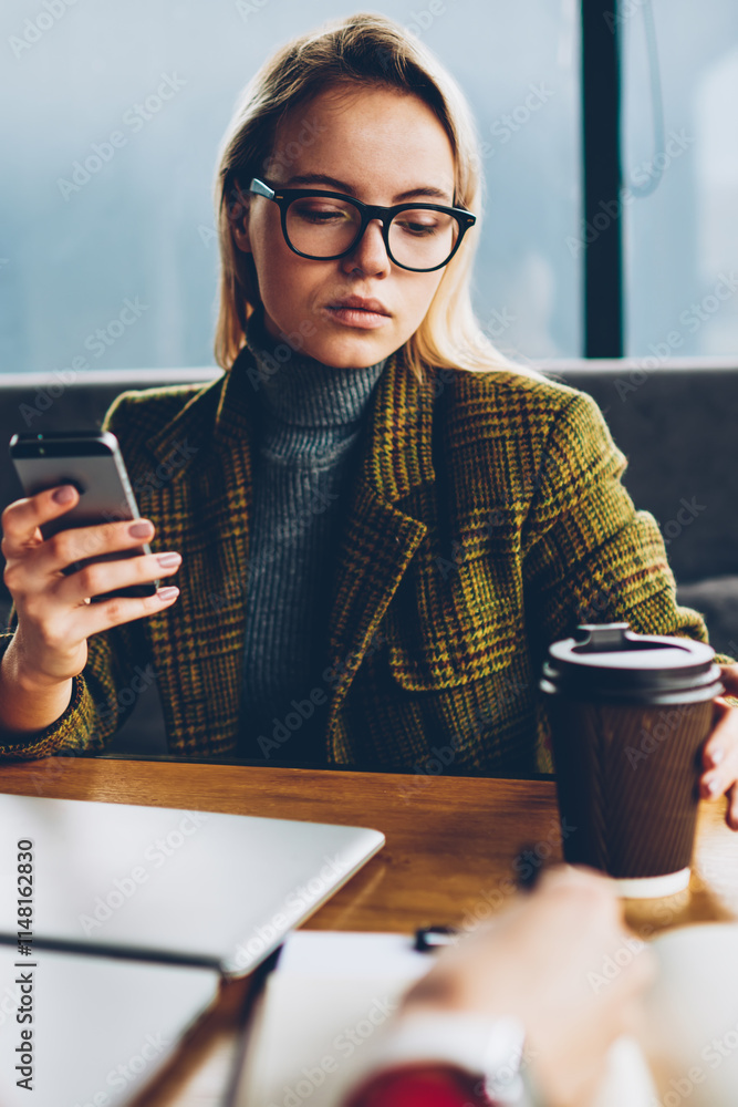 Poster serious female blogger dressed in elegant jacket taking coffe to go in hand sitting at table and che