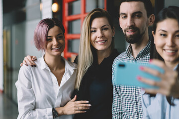 Diversity group of emotional colleagues dressed in casual outfit making selfie on smartphone camera while standing indoors.Smiling man and women having fun during taking together picture on phone