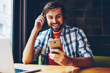 Cheerful bearded hipster guy choosing song in music app to listen in modern headphones connected...