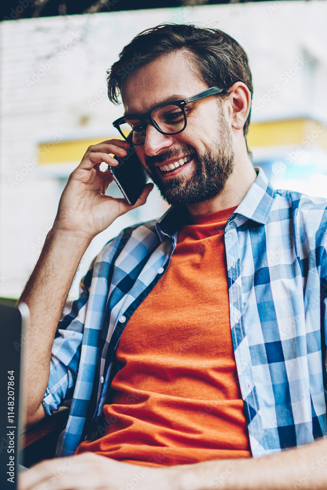 Poster happy bearded young man in eyeglasses communicating on smartphone while updating software on modern 