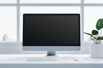 Fototapeta premium Modern desktop computer on a white desk near a plant, with large windows in the background, in a bright office setting. 3D Rendering