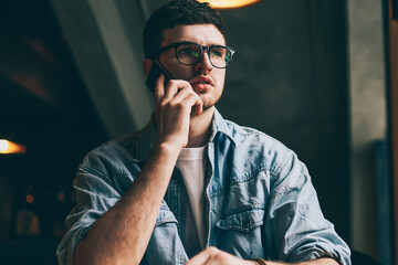 Serious concentrated man calling to operator for checking account balance using smartphone with 4g internet, pondering hipster guy in spectacles making telephone conversation with colleague indoors