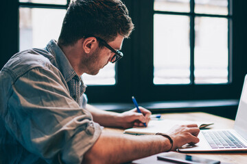 Young millennial male student searching information for homework using laptop and wifi connection indoors, man writing article in textbook for add interesting content on own website or web page