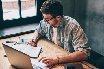 Hipster guy pondering on idea for essay sitting at wooden table with laptop device and looking on...