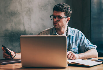 Handsome male it professional checking time on smartphone device and updating notification indoors, casual hipster guy sitting in front of laptop and dialing number on cellular phone via app