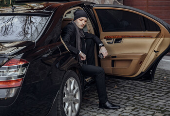 A man in a black suit and hat is getting into a black car. The car is parked on a street with a...
