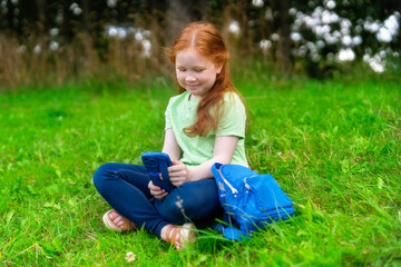 Fototapeta premium Young girl sits on green grass interacting with her smartphone in a sunny outdoor setting