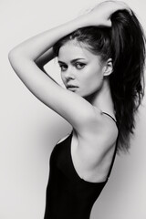 Confident young model posing in a sleek black swimsuit, gracefully pulling her hair up, showcasing a strong silhouette on a minimalist white background.