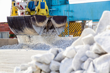Loading at the port. The large grab of a gantry crane dumps white marble chips onto an open warehouse on the wharf