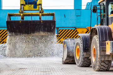 White marble crumb spills from the grab of a gantry crane in front of a yellow bucket truck