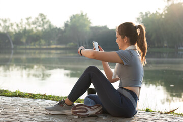 Fototapeta premium Young Woman Sitting and Resting After Exercising by a Lake, Checking Her Smartphone, Enjoying Nature and Relaxing Outdoors