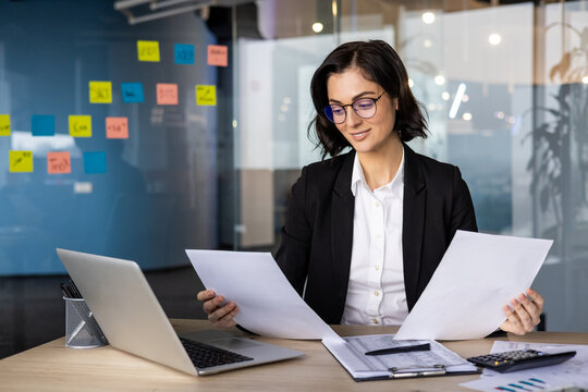 Professional businesswoman reviewing documents at desk with laptop in modern office space. Reflects focus, productivity, and confidence in corporate setting. Concept of work and strategy.
