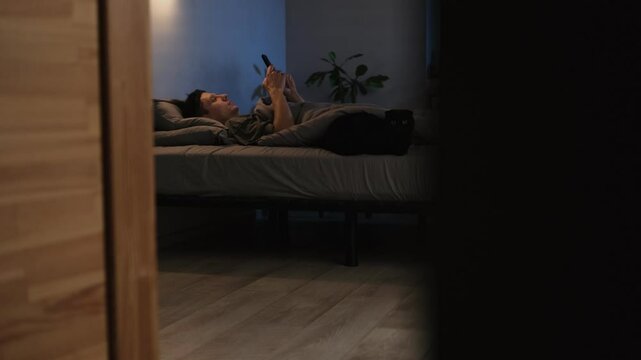 Man relaxing in bed at night, scrolling through his phone to unwind after work. People consume content from social media. The cat is lying next to him