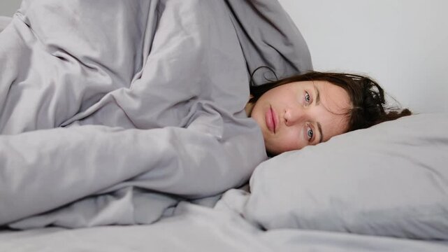 Young depressed woman hiding under a blanket in a gray, minimalist room, struggling with depression and emotional distress.