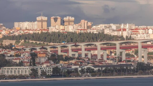 Aerial timelapse of Lisbon skyline featuring colorful houses, Amoreiras shopping center towers and the 25th of April Bridge. Historic district with green trees and Prazeres Cemetery in the foreground