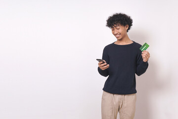 young man standing wearing blue sweater, holding phone, credit card and looking at phone with smile isolated on white background.
