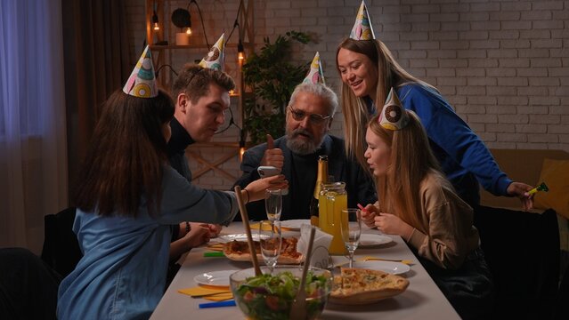 Family sit at table at home, grandchildren and parents celebrating birthday of grandfather, family members showing grandpa selfie on smartphone.