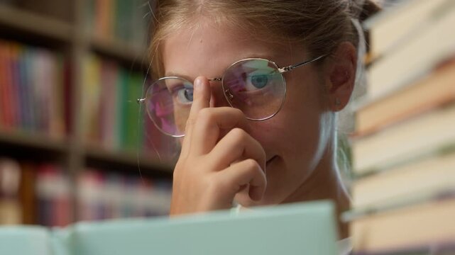 Close up clever smart intelligent Caucasian little girl playful schoolgirl child school kid pupil learner student in glasses eyeglass looking at camera reading book literature inside library education