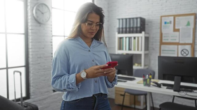 Woman standing in a modern office looking at her smartphone with a focused expression, surrounded by desks and office supplies, large windows and shelves in the background.