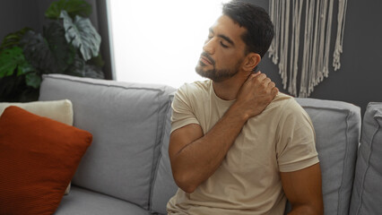 Obraz premium Young man with beard sitting on sofa holding neck in pain indoors, showing discomfort in modern living room environment.