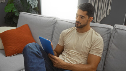 Young hispanic man with beard relaxing in living room using tablet showing modern comfortable home interior design.