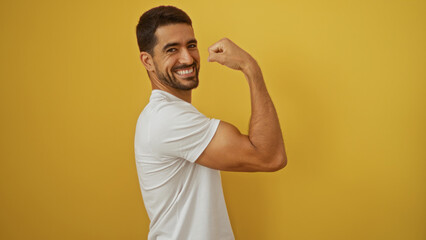 Young hispanic man flexing muscle smiling confidently against a vibrant yellow background showcasing strength and positivity in a casual white t-shirt setting.