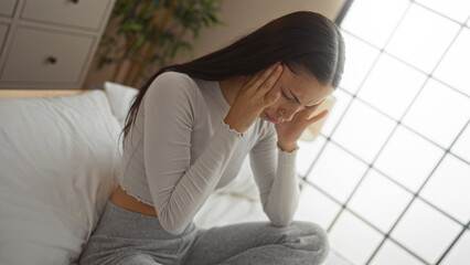 Woman sitting on bed in bedroom holding her head with a pained expression suggesting headache or...