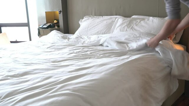 A person is pulling a white pillow off of a bed. The bed is unmade and has a white comforter