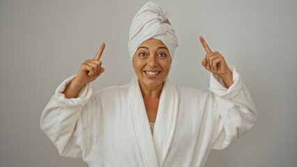 Mature woman smiling in white bathrobe and towel on head pointing fingers upward against white background