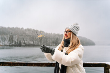 young woman holding a sparkling sparkler and smiling happily in a serene winter landscape by a...