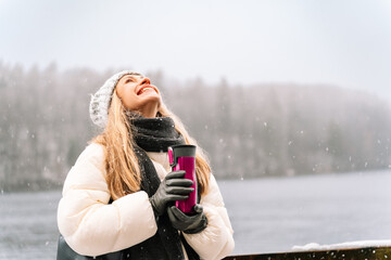 smiling woman enjoying snowfall with a thermos in winter nature