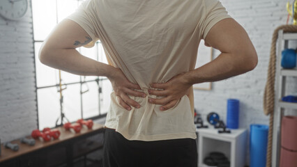 A man holds his back in pain while standing in a gym, surrounded by weights, ropes, and fitness...