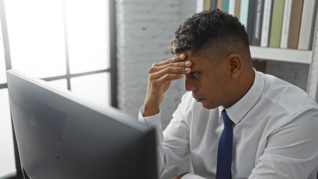 Young man sitting in an office looking at his computer screen with a thoughtful expression, dressed in professional attire, surrounded by bookshelves and large windows