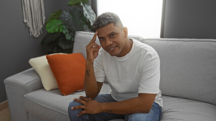 Young hispanic man sitting on a grey sofa, smiling in a modern living room with green plants and pillows, showcasing a relaxed indoor home environment.