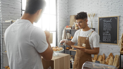 Men working and interacting at a bakery, with one man serving fresh pastries to a customer in a cozy indoor shop setting with breads and baked goods displayed in the background.