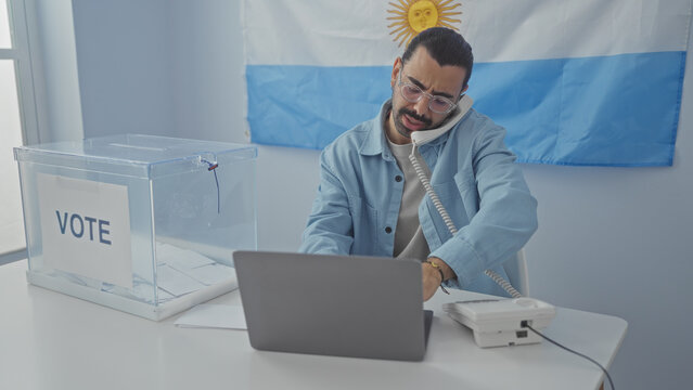 Young man with moustache in indoor voting room in argentina using laptop and phone near ballot box and argentine flag