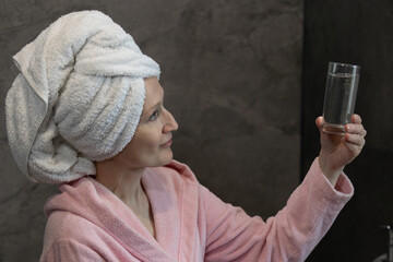 Portrait of a beautiful young woman in pink bathrobe and towel on head, in the bathroom, holding glass of water