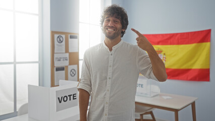 Young man smiling and pointing at himself inside an electoral room with spanish flag and voting...