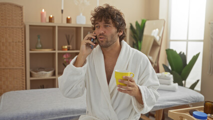 Young man talking on phone while holding a yellow mug in a spa room with candles and plants creating a serene wellness center ambiance