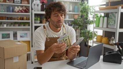 Young man counting dollar bills in a home decor store while sitting at a desk with a laptop and cardboard box