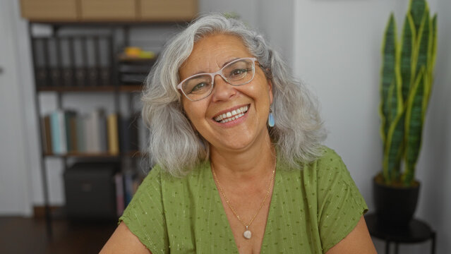 Elderly woman smiling warmly in an indoor office setting, with grey hair and glasses, serving as a mentor or professional in a serene workplace environment, surrounded by books and plants.