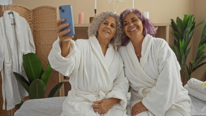 Middle-aged women wearing white robes sit together smiling and taking a selfie in a spa room with plants and soft lighting, capturing a moment of relaxation and friendship.